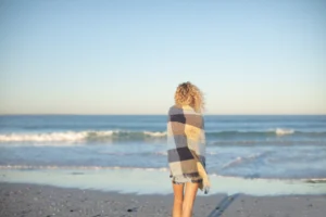 Mulher de costas envolta em manta observando o mar em uma praia no inverno, com céu limpo e ondas suaves