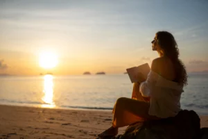 Mulher lendo à beira-mar ao pôr do sol, refletindo sobre bem-estar e saúde mental com apoio de psicólogo online